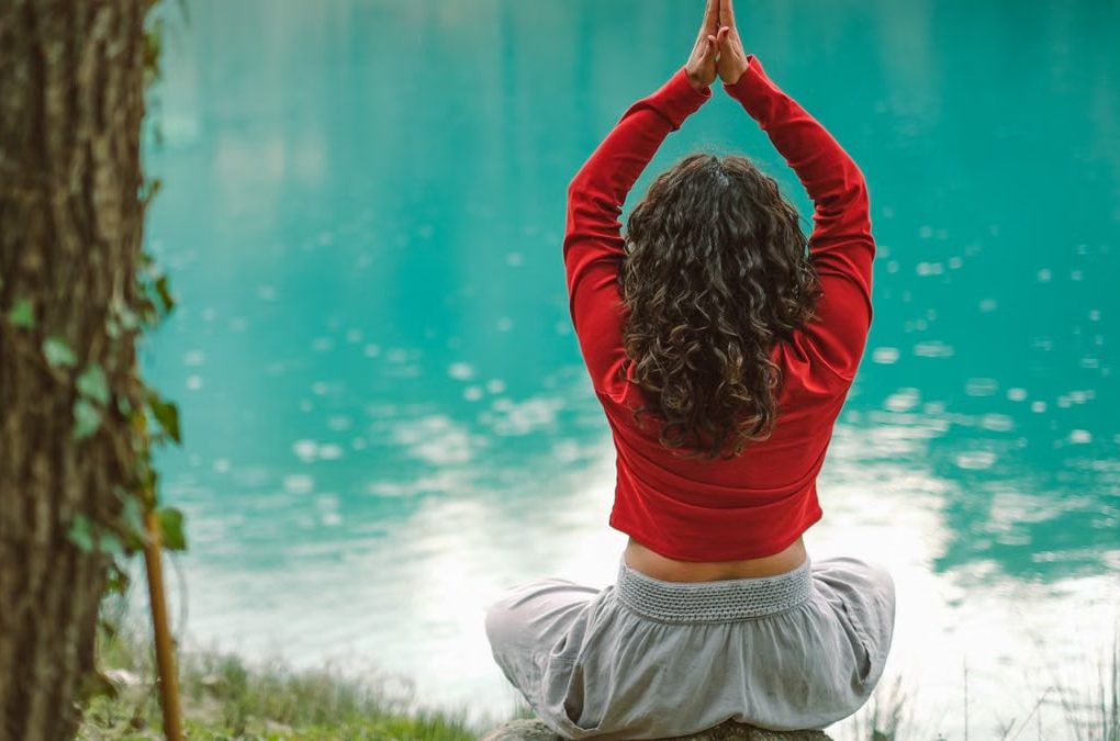 A woman meditates in nature on a rock by a tranquil turquoise lake, practicing yoga.
