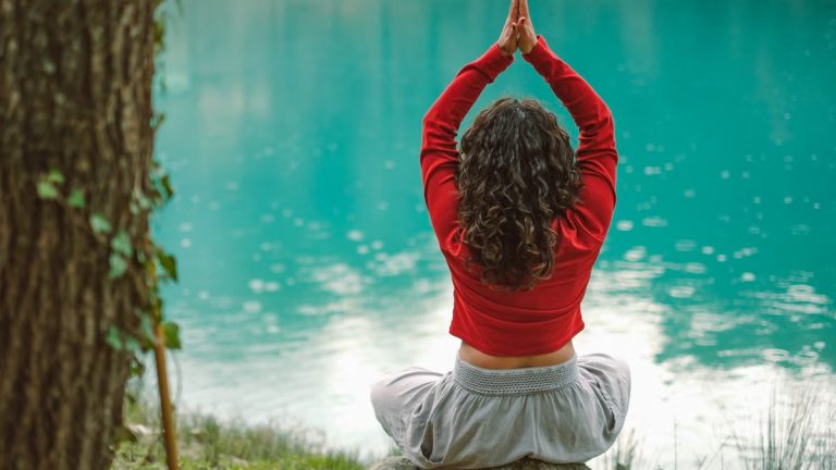 A woman meditates in nature on a rock by a tranquil turquoise lake, practicing yoga.