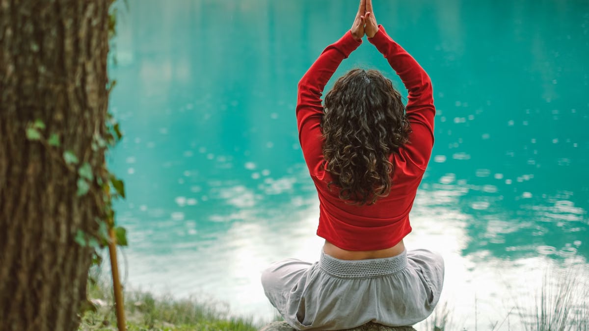 A woman meditates in nature on a rock by a tranquil turquoise lake, practicing yoga.